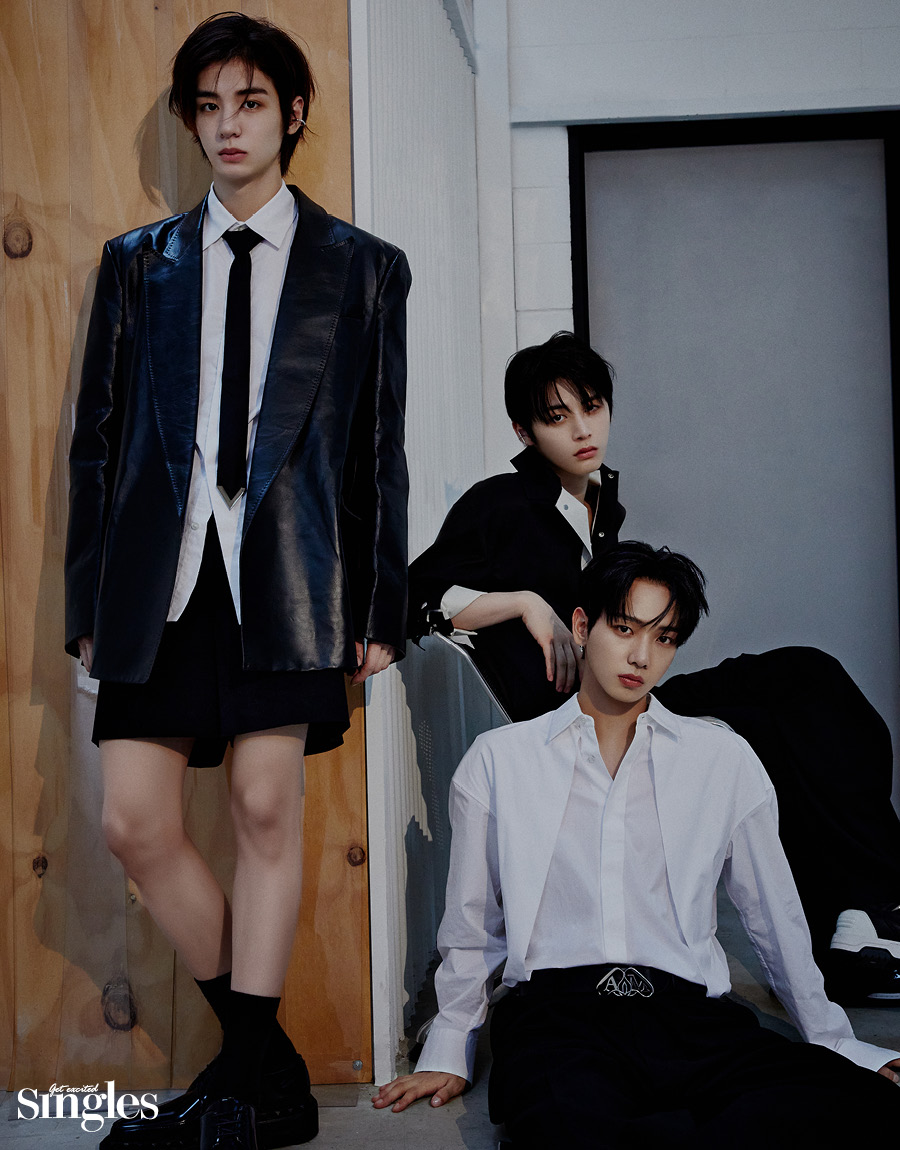 보이넥스트도어 멤버 이한이 벽에 기대어 서 있고, 재현과 태산이 그 옆에 앉아 정면을 바라보고 있다.